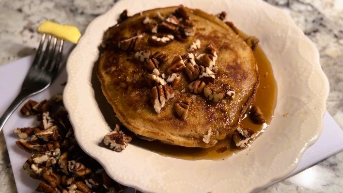pancakes covered in maple syrup and pecans on white plate beside butter and pecans