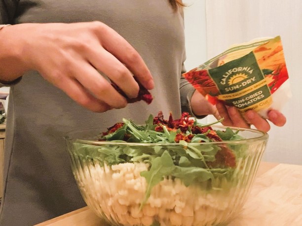 lady putting sundried tomatoes into salad in a glass bowl
