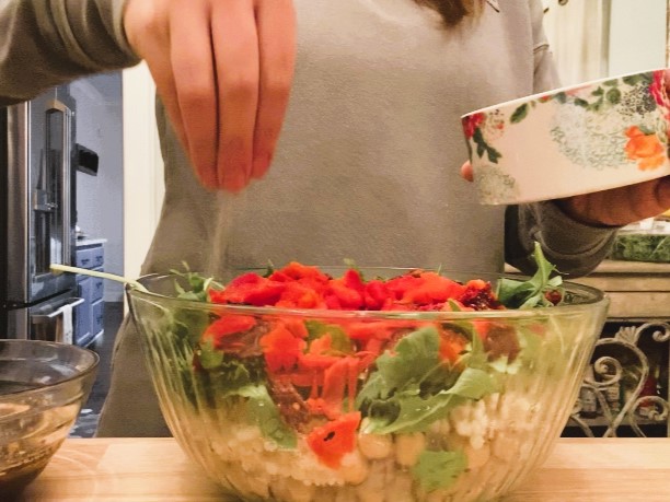 woman sprinkling salt onto Mediterranean couscous salad in a glass bowl