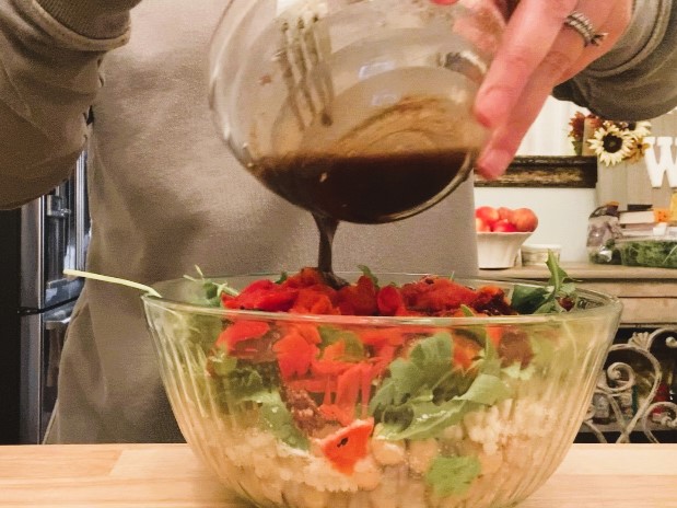 woman pouring balsamic vinaigrette on top of Mediterranean couscous salad in glass bowl
